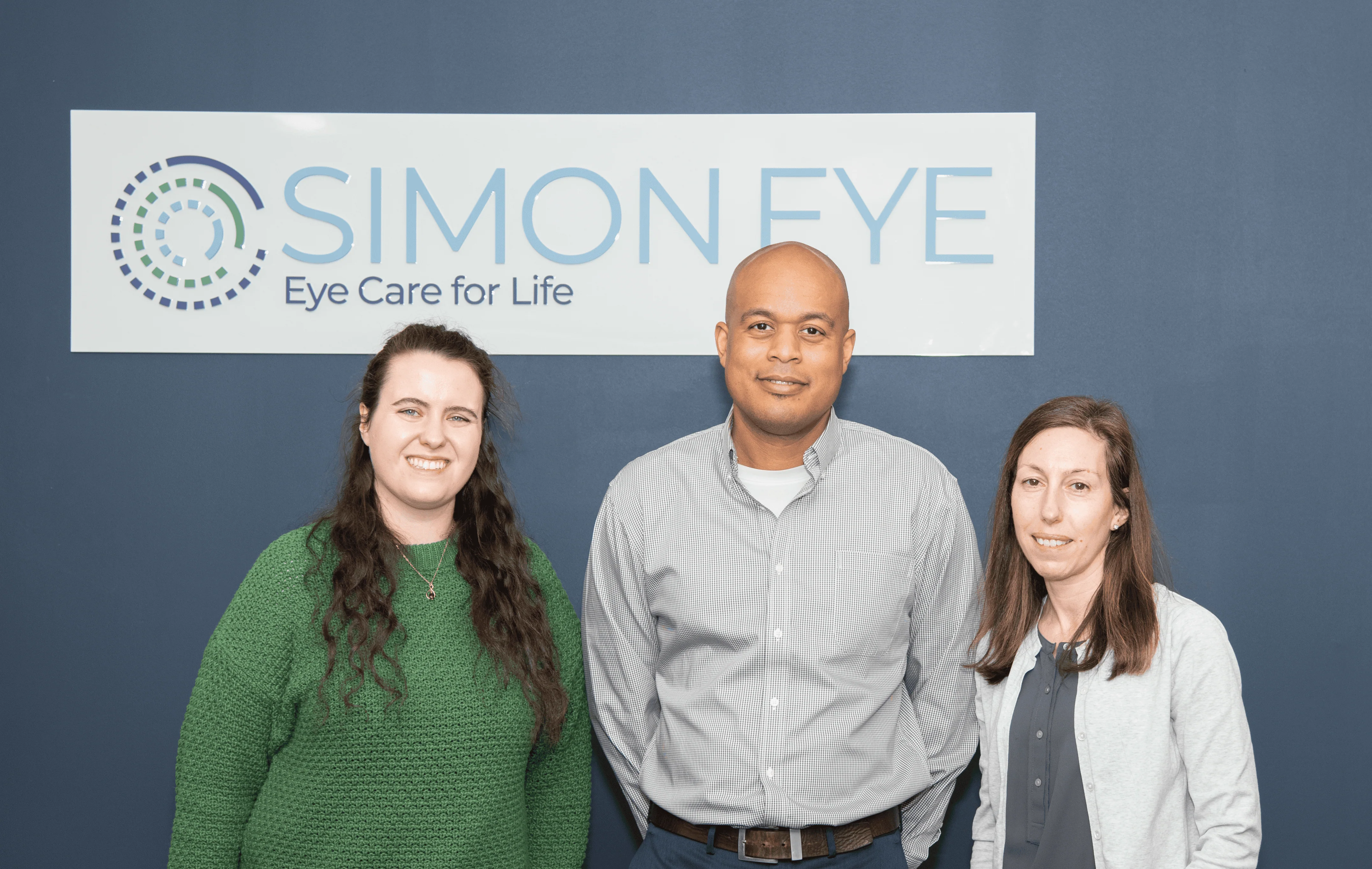 Three people standing in front of a sign that reads "Simon Eye: Eye Care for Life." They are all facing the camera and smiling.